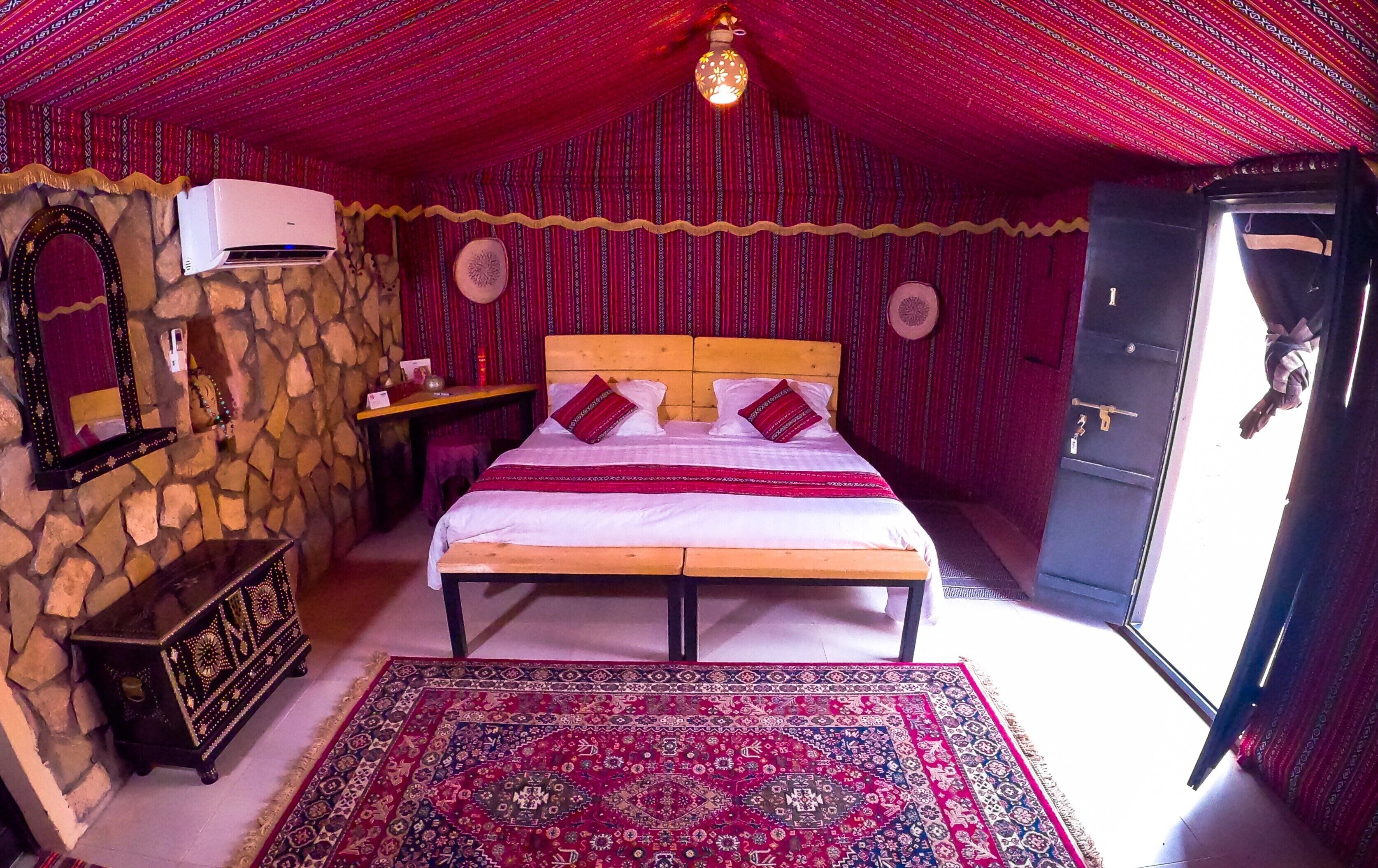 A room with a stone wall, a bed with red and white bedding, a wooden headboard, a mirror, a small table with a lamp, and a patterned rug.