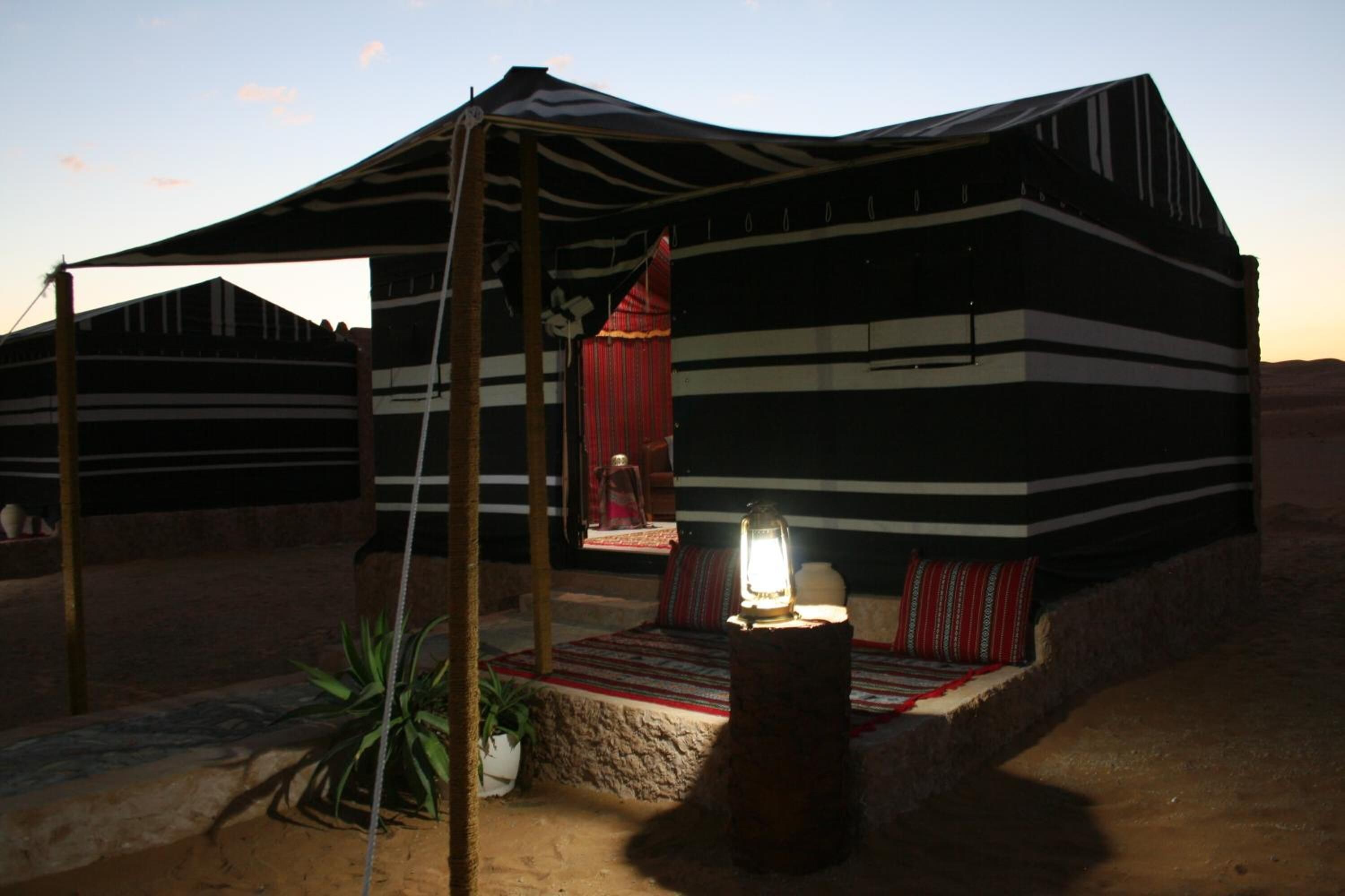 Una tienda de campaña en el desierto con exterior a rayas, un pequeño porche con cortina roja y un farol en el suelo.
