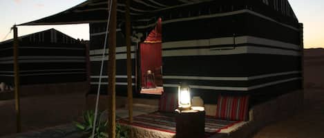 Una tienda de campaña en el desierto con exterior a rayas, un pequeño porche con cortina roja y un farol en el suelo.