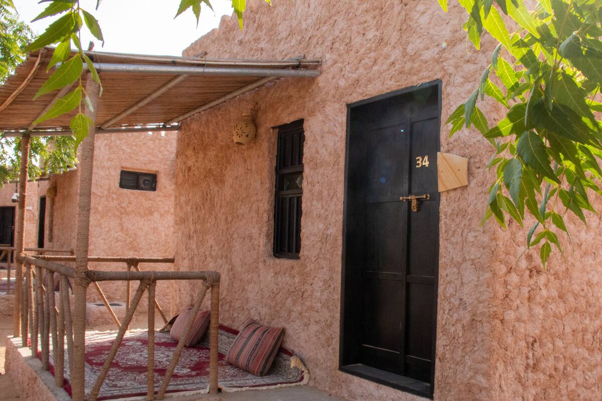A rustic building with a black door marked with the number 34, a small window, and a covered porch area with a patterned rug and cushions.