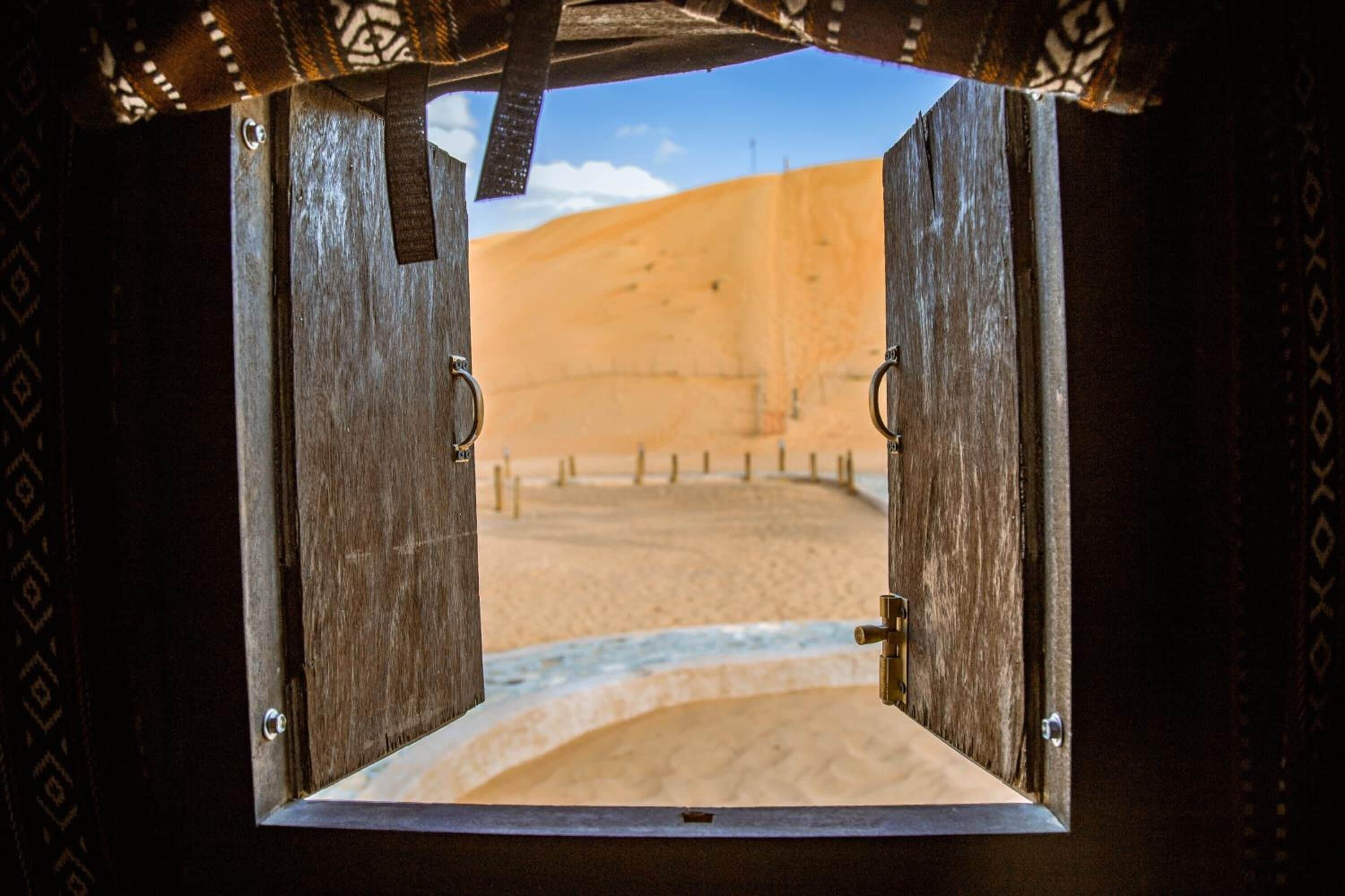 Une porte en bois, dotée de poignées en métal, qui donne sur un paysage désertique avec des dunes de sable et une clôture.