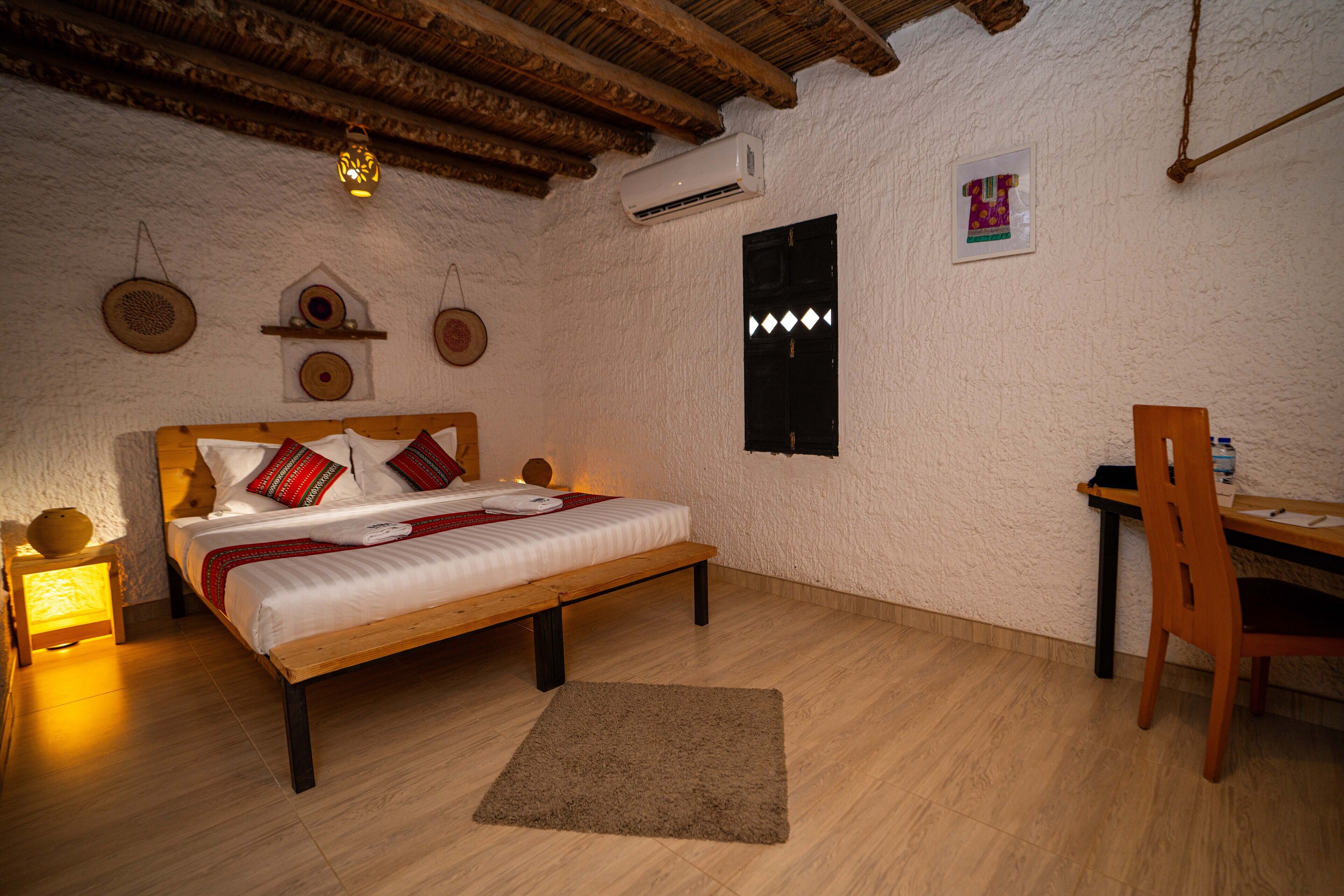 Una habitación con una cama de madera, un escritorio y una silla. Tiene un aire acondicionado de pared y una mesita con una lámpara.