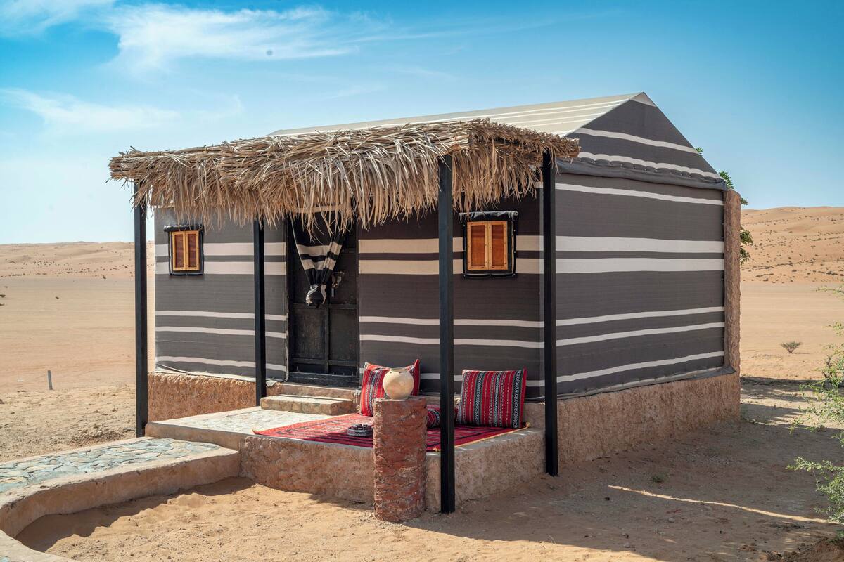 A desert-style building with a striped exterior, a thatched roof, and a small porch with cushions.
