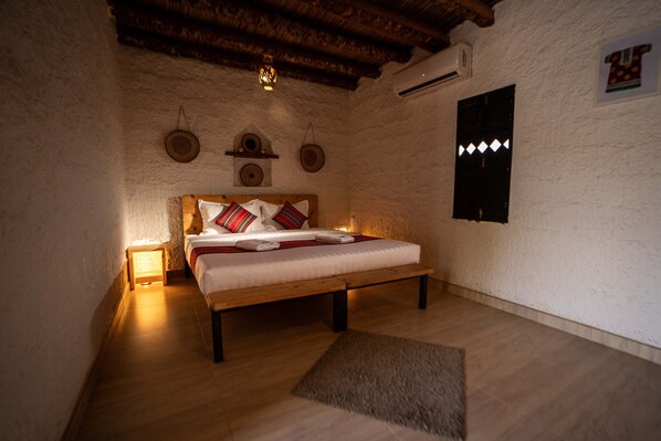 A cozy bedroom with a wooden bed, a small bedside table with a lamp, a wall-mounted shelf with decorative items, and a wooden floor.