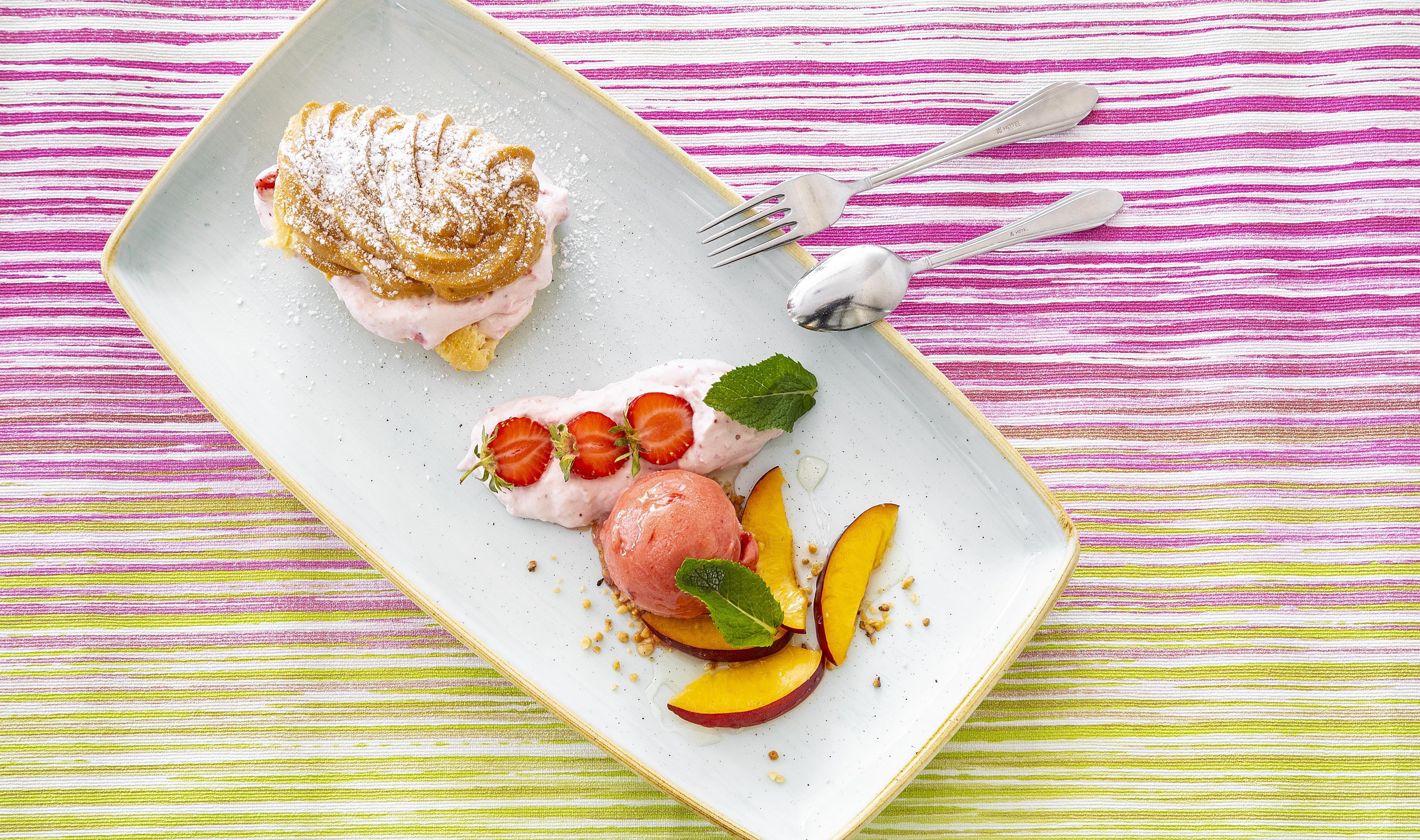 Un plato de postre con un pastel, helado y rodajas de fruta sobre un mantel a rayas.