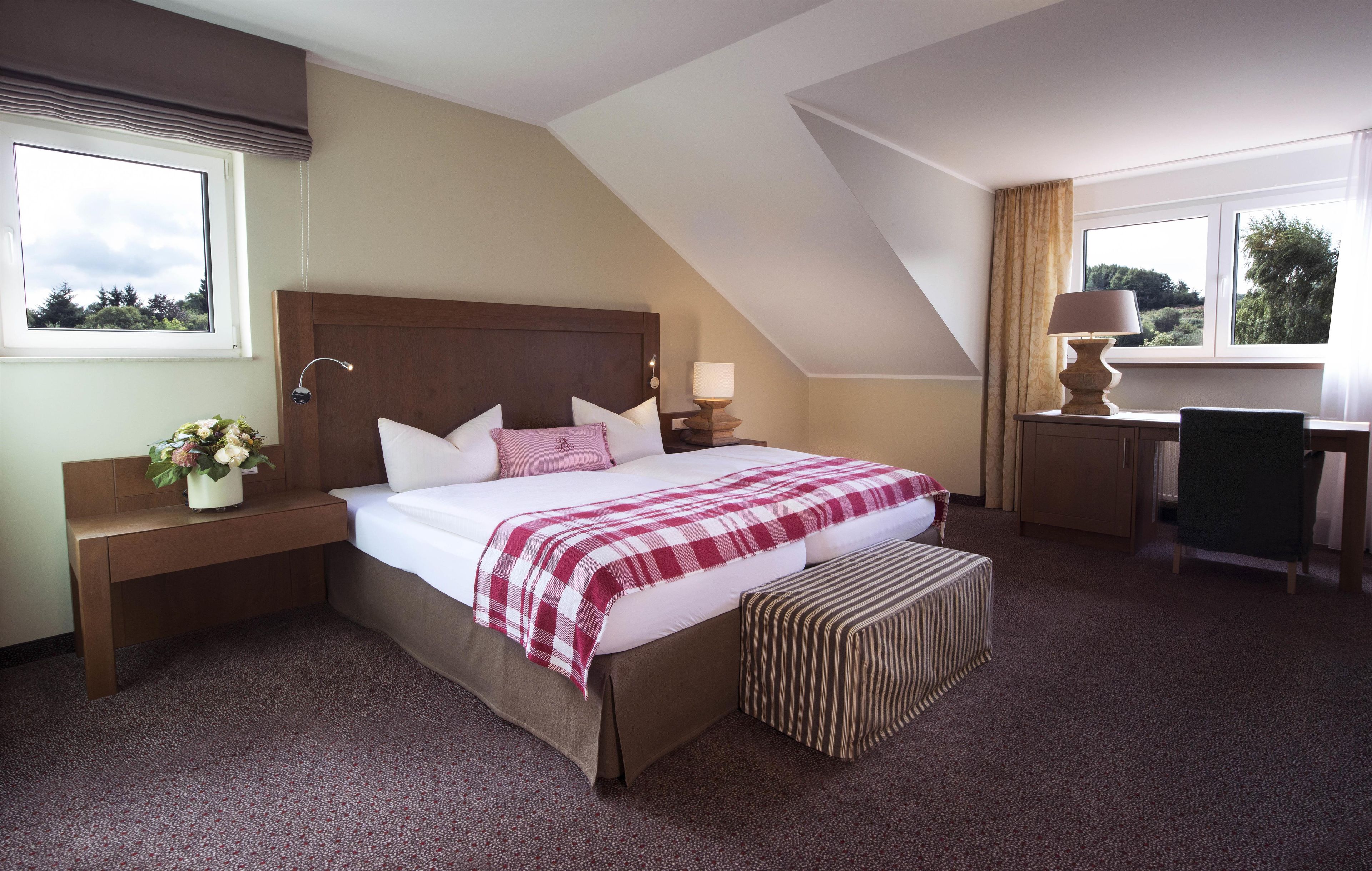 A hotel room with a large bed, a desk, and a chair. There is a window with curtains and a view of trees.