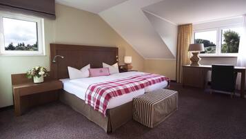 A hotel room with a large bed, a desk, and a chair. There is a window with curtains and a view of trees.