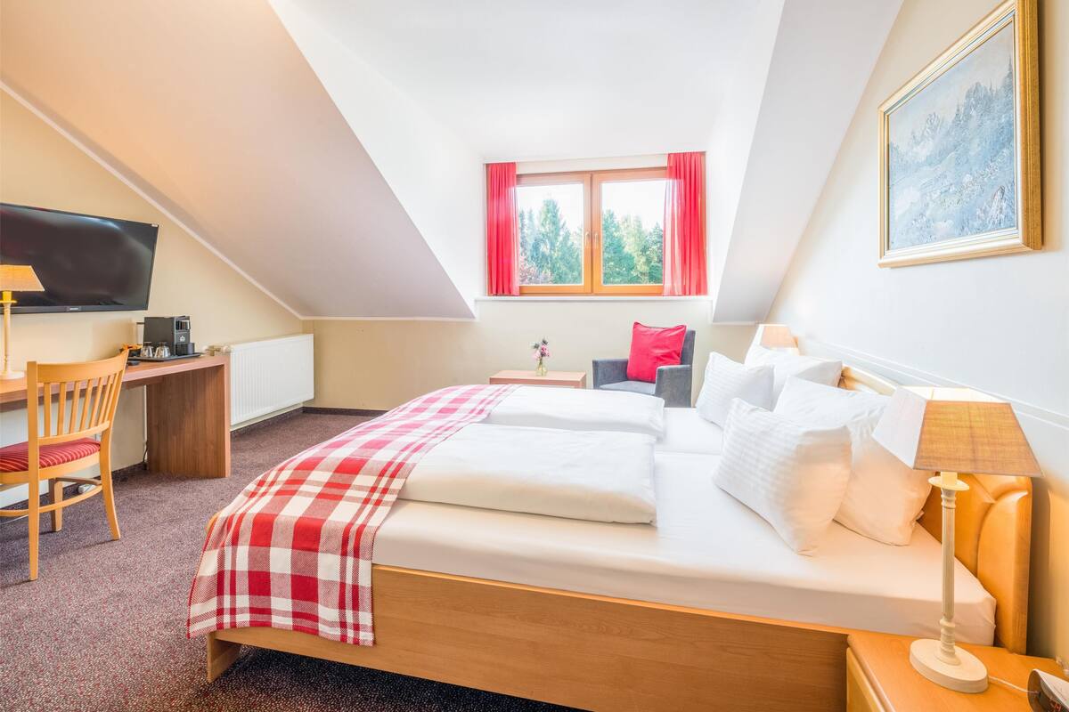 A bedroom with a bed, a desk, a chair, a TV, and a window with curtains.