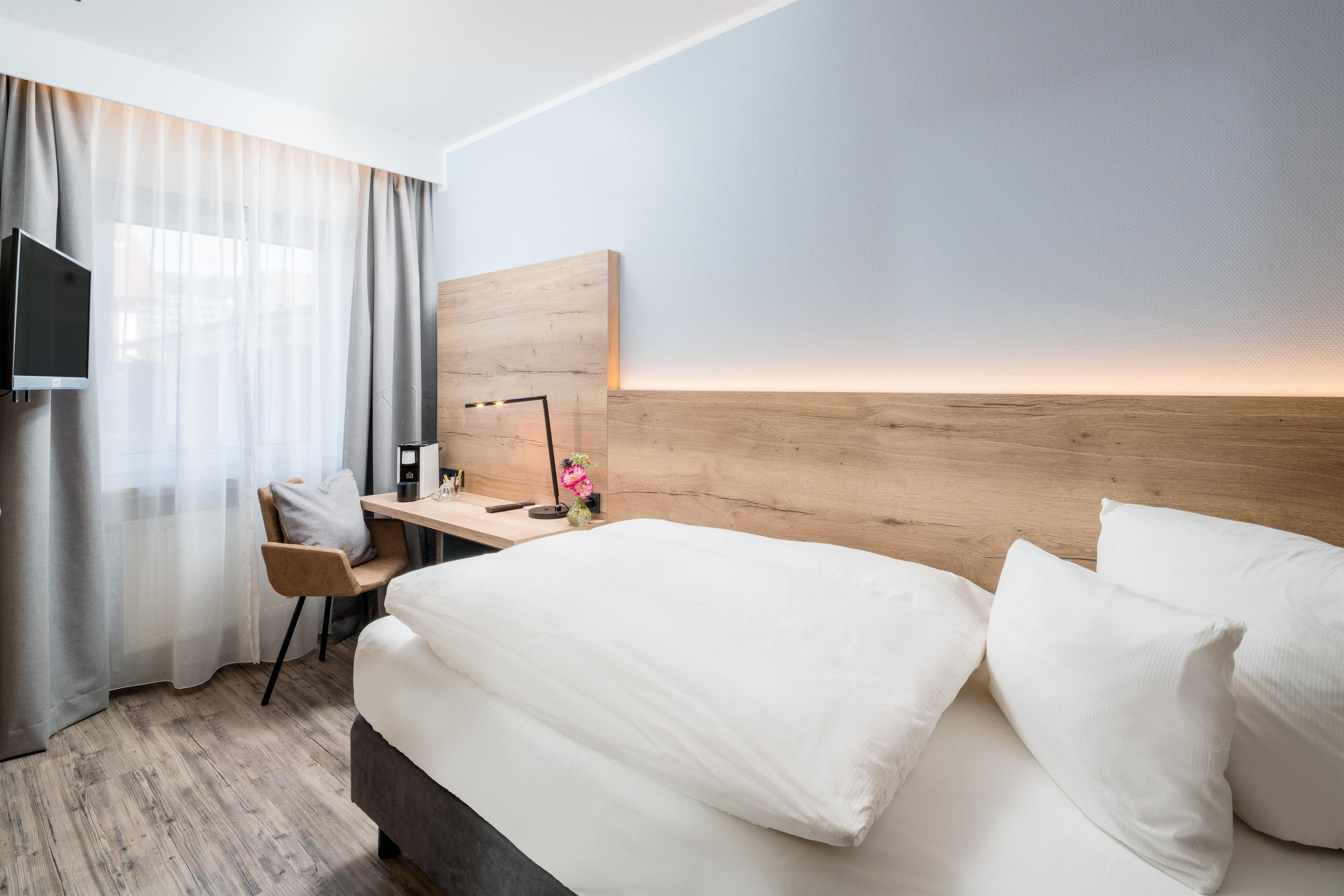 Habitación de hotel con cama, escritorio, silla, televisor y mesita de noche con lámpara y flores.