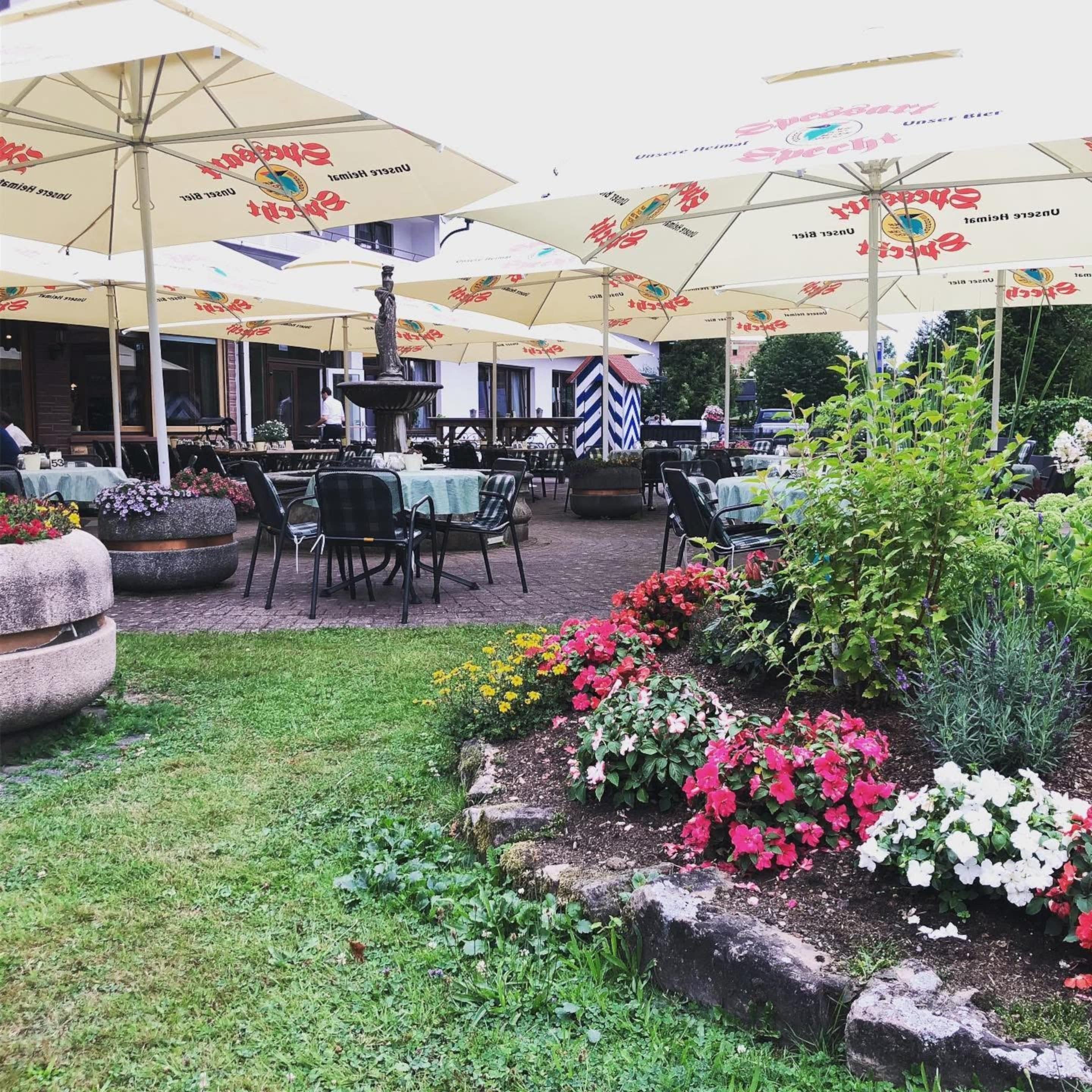 An outdoor dining area with tables and chairs, umbrellas, and a garden with flowers and plants.