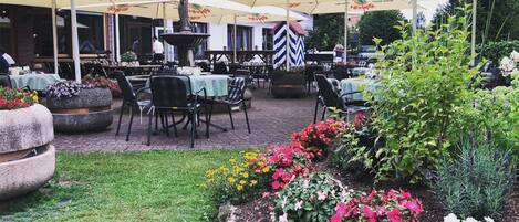 An outdoor dining area with tables and chairs, umbrellas, and a garden with flowers and plants.