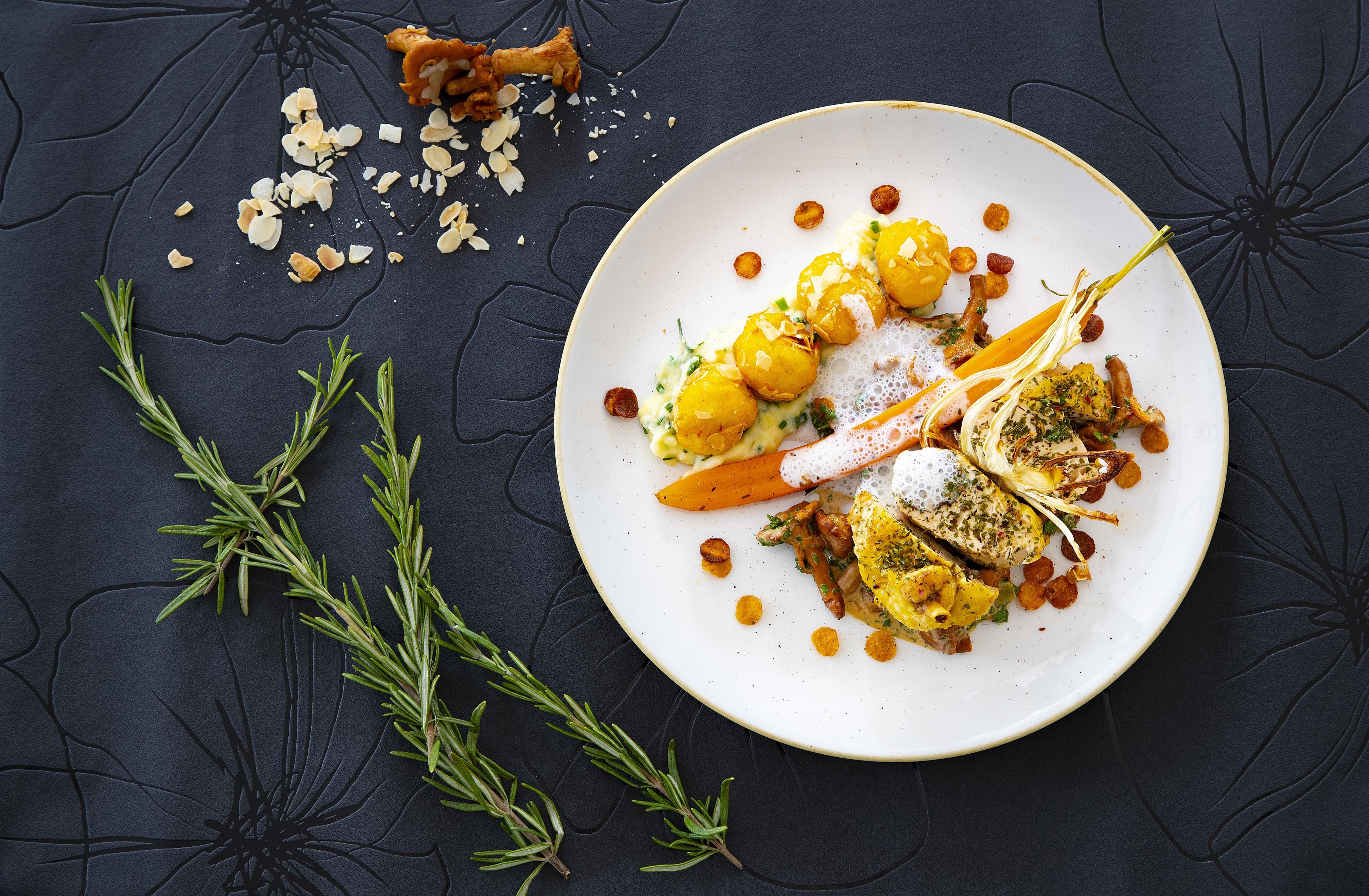 Un piatto di cibo con diversi elementi, tra cui verdure arrostite, noci e erbe aromatiche, su una superficie scura e strutturata.