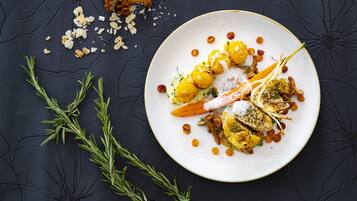 Un piatto di cibo con diversi elementi, tra cui verdure arrostite, noci e erbe aromatiche, su una superficie scura e strutturata.