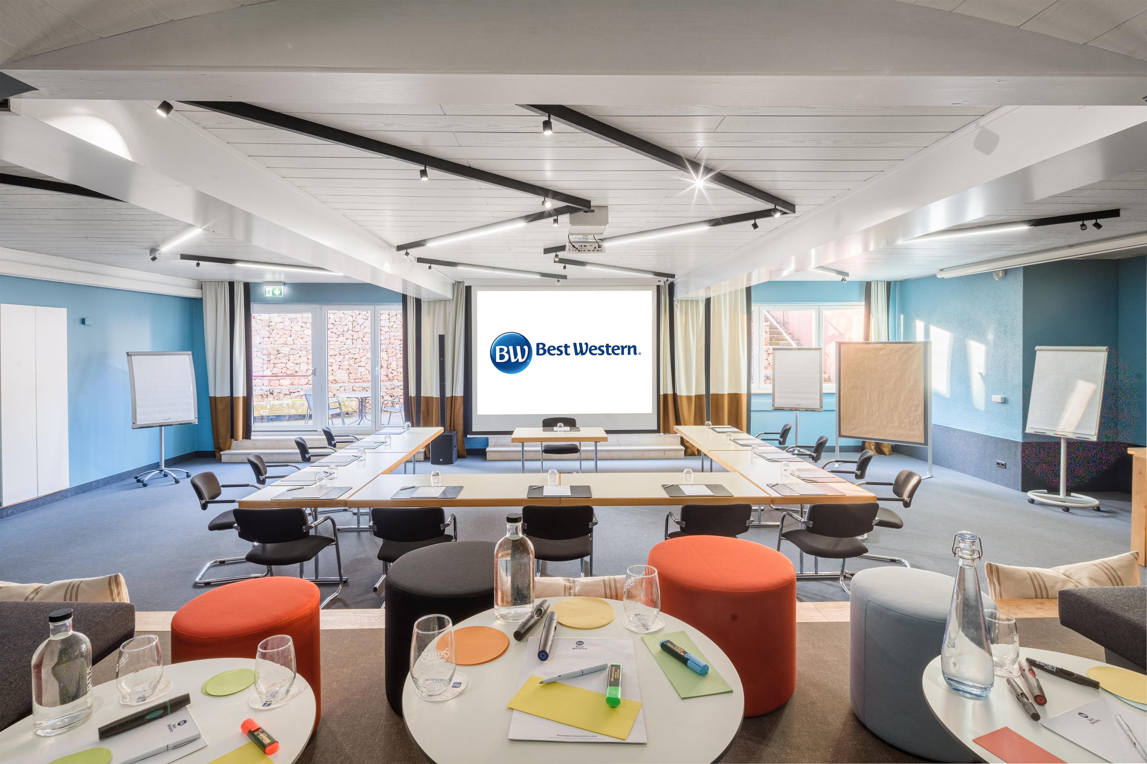 A conference room with a large screen displaying 'Best Western' branding, multiple tables with chairs, and a casual seating area with ottomans and a water bottle.