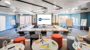 A conference room with a large screen displaying 'Best Western' branding, multiple tables with chairs, and a casual seating area with ottomans and a water bottle.
