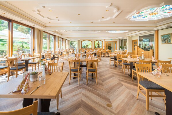 A spacious restaurant with wooden tables and chairs, large windows, and a decorative ceiling.