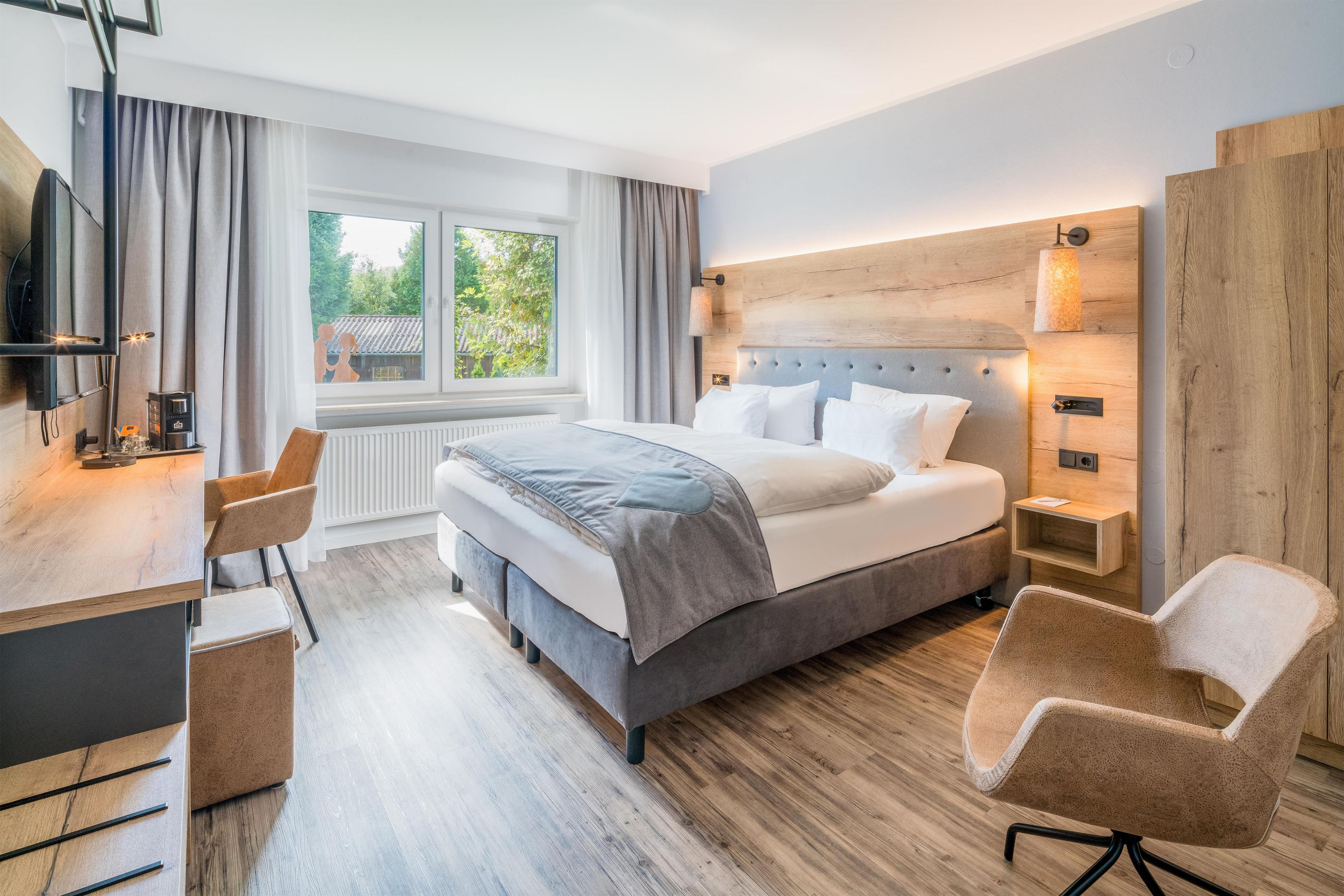 Habitación de hotel moderna con una cama grande, un escritorio, dos sillas, una ventana con cortinas y vistas a zonas verdes.