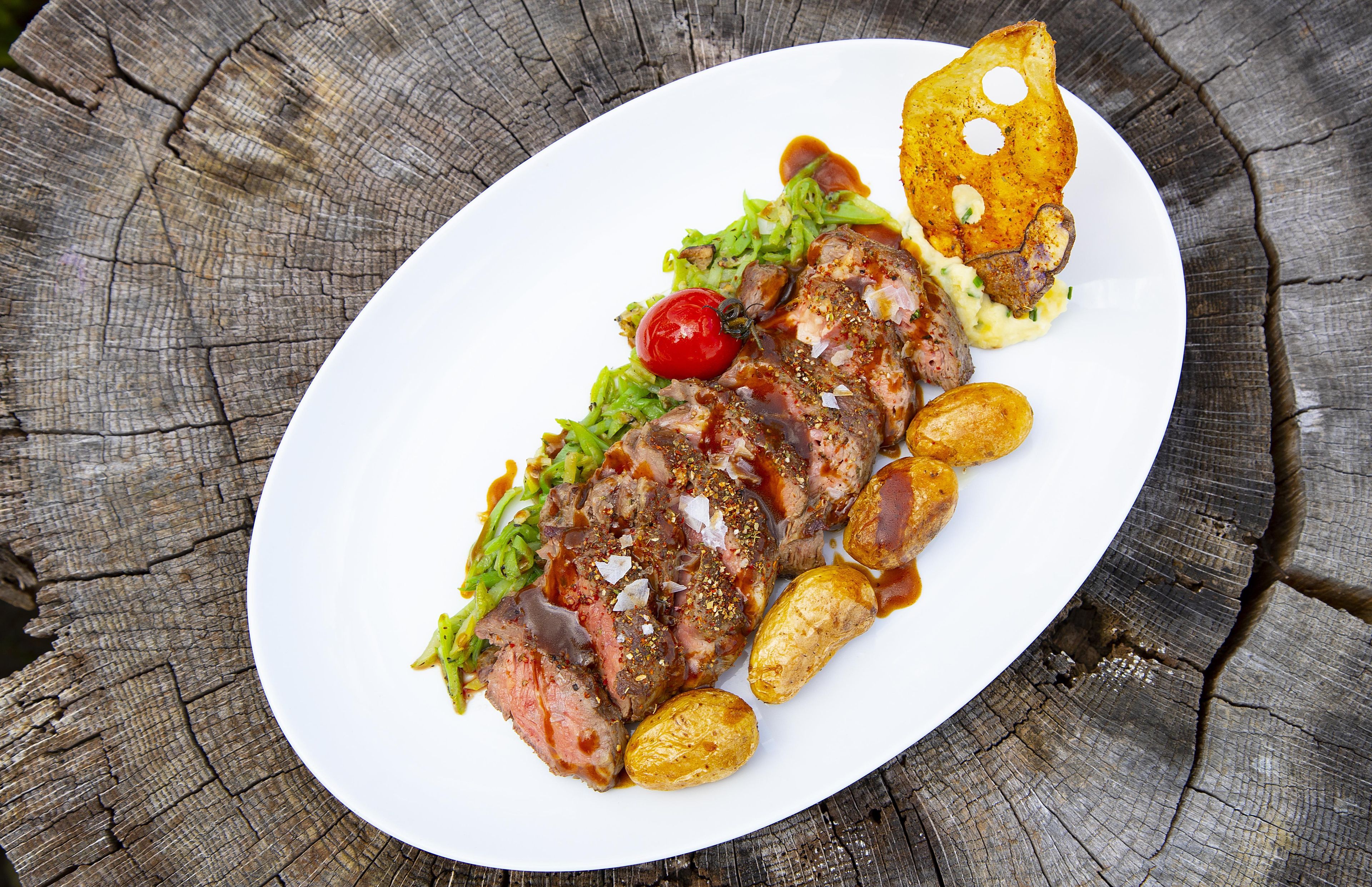 Une assiette de nourriture composée de viande, de pommes de terre et d’une tomate cerise, le tout servi dans une assiette blanche.