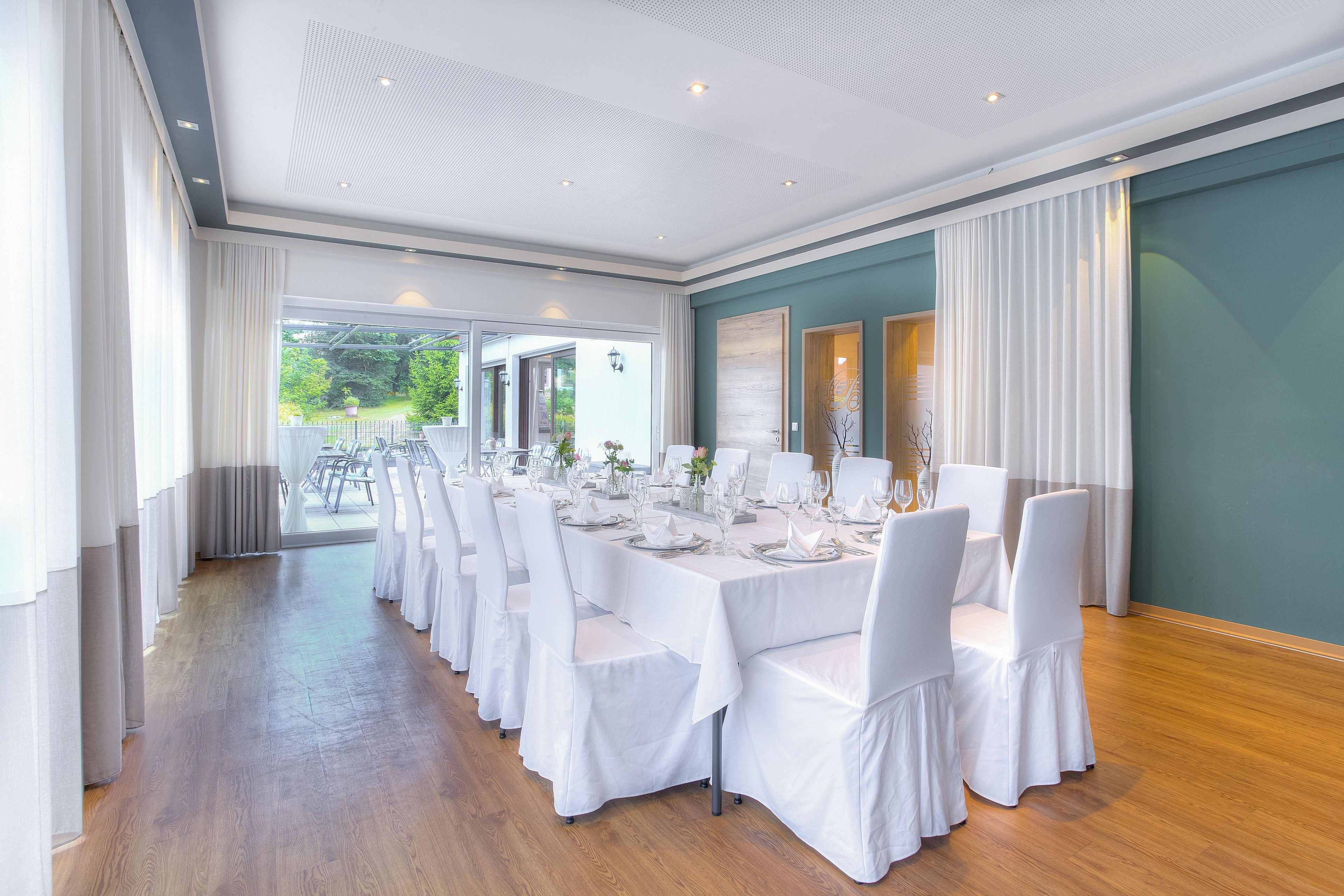 A long dining table set for a formal event, with white tablecloths and chairs, in a room with a wooden floor and a view of greenery outside.