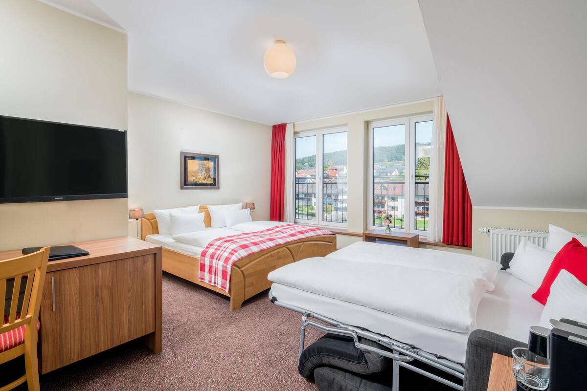 A hotel room with two beds, a desk, a chair, a TV, and large windows with red curtains.