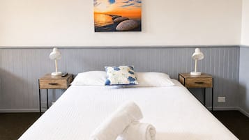 A hotel room with a bed, two bedside tables with lamps, a pillow, and a painting of a sunset over rocks.