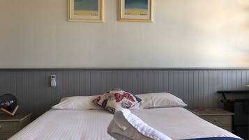 Une chambre d’hôtel avec un lit, deux œuvres d’art encadrées accrochées au mur, une table de chevet avec une lampe et une petite table de nuit avec un livre et une horloge.