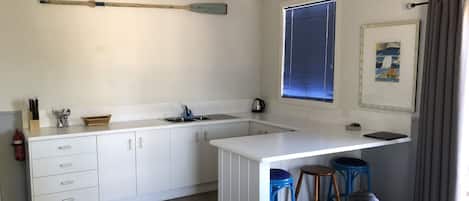 A modern kitchen with white cabinets, a large window with blinds, a wooden paddle on the wall, and a blue stool.