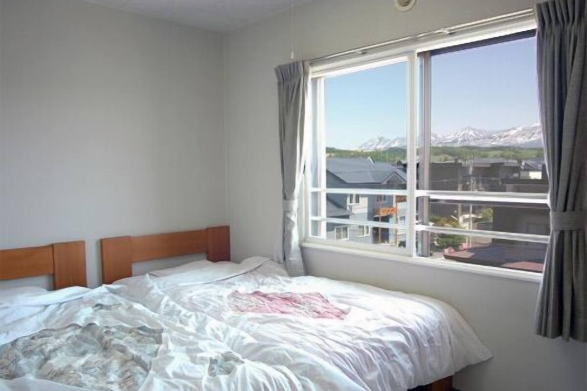 A bedroom with a large window, a bed with a white duvet, and a view of buildings and mountains.