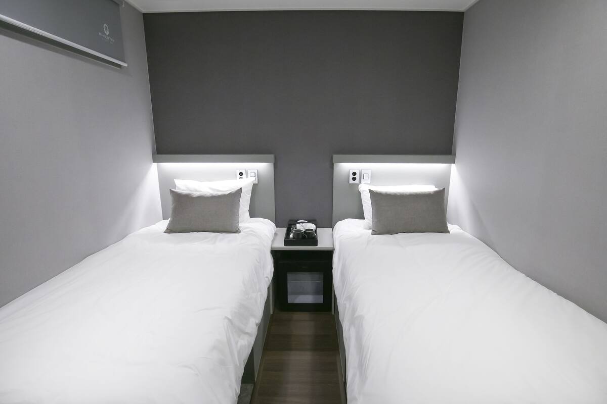 Habitación de hotel con dos camas individuales, cada una con cabecera, una mesita con bandeja y una mesita de noche.