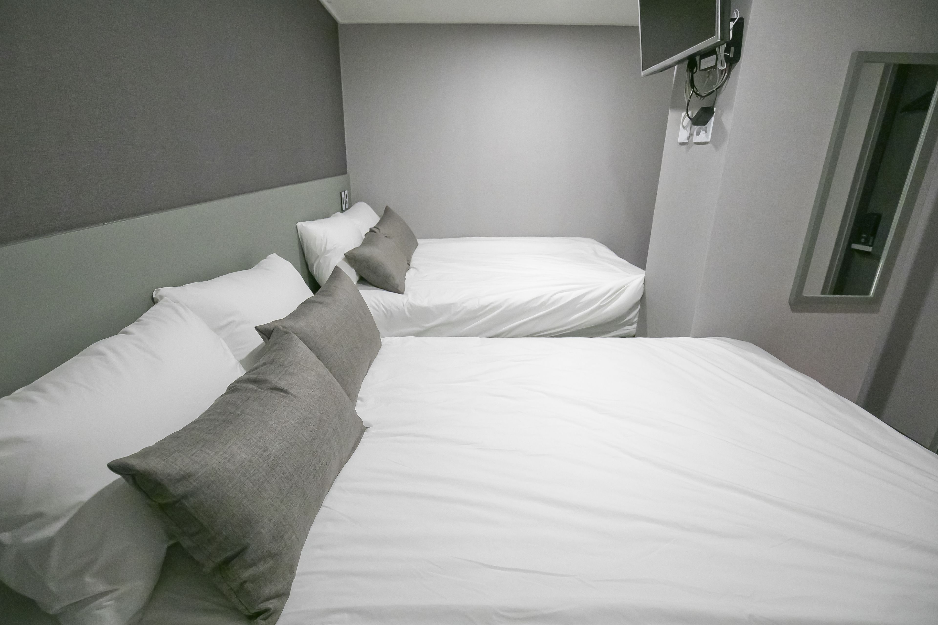 A twin room with two single beds, each with white linens and grey pillows. A small bedside table with a lamp is visible on the left bed. The room has a neutral color scheme with grey walls and a white ceiling.