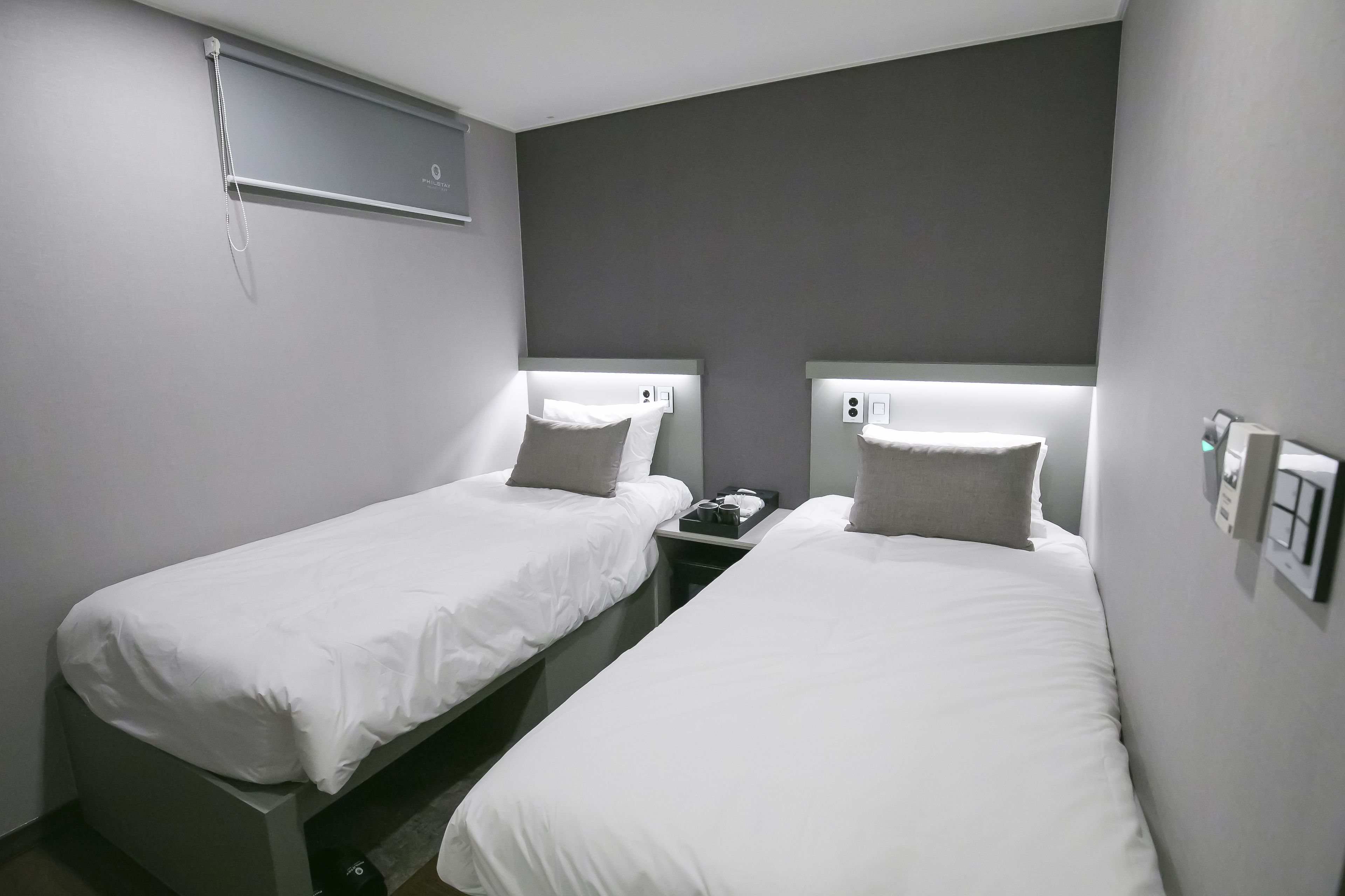 Una habitación de hotel con dos camas individuales, cada una con cabecera y almohada. Hay una mesita de noche con un teléfono y una pequeña mesa entre las camas.