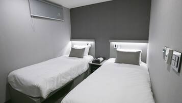 Una habitación de hotel con dos camas individuales, cada una con cabecera y almohada. Hay una mesita de noche con un teléfono y una pequeña mesa entre las camas.