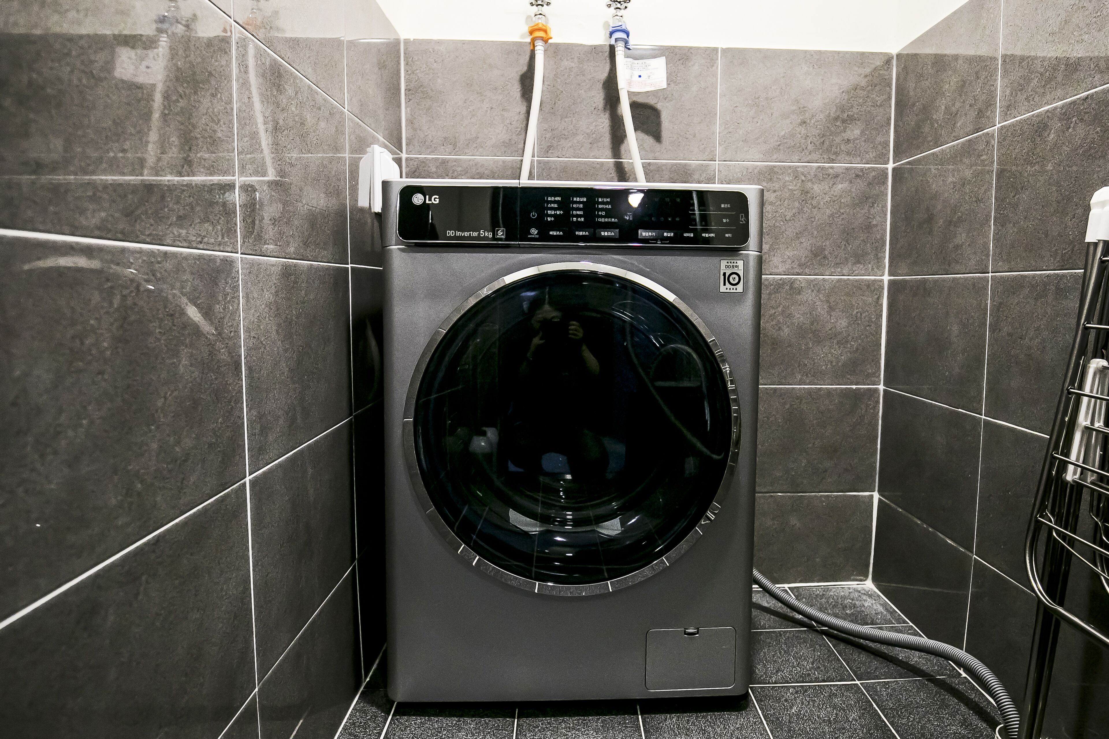 Un lave-linge LG moderne et compact, d’une capacité de 5 kg, à chargement frontal, placé dans un coin d’une pièce dont les murs sont recouverts de carreaux gris foncé et où se trouve une étagère métallique sur la droite.