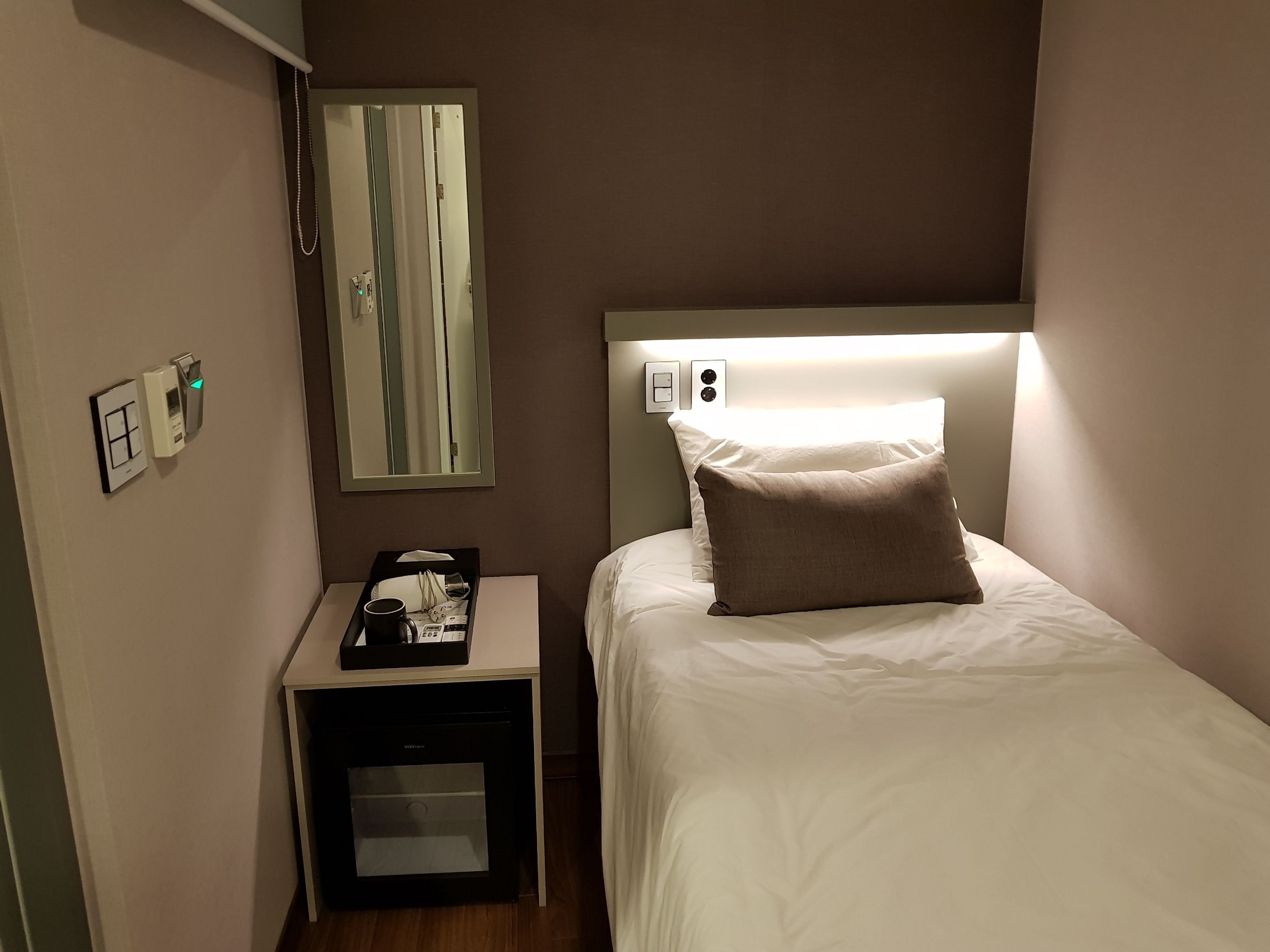 A hotel room with a bed, a nightstand, a mirror, a wall-mounted light, and a small cabinet.