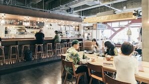 Un café moderne avec des tabourets et des tables en bois, un bar doté d’un comptoir et de tabourets, et un tableau des menus bien visible.