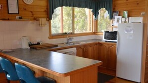 A compact kitchen with wooden cabinets, a sink, a microwave, and a refrigerator. There are two blue bar stools and a window with curtains. - Coles Bay House (Coles Bay)