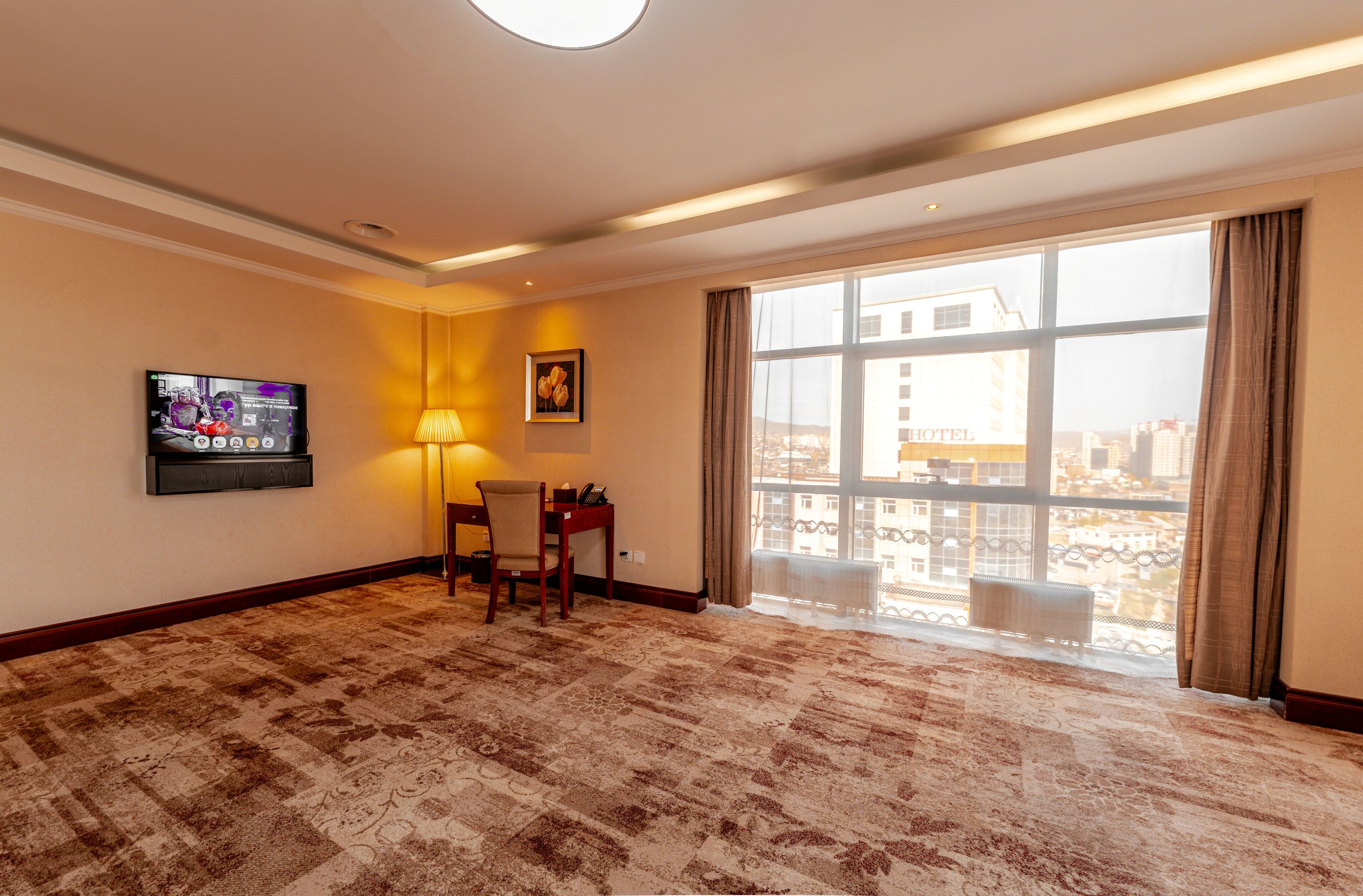 A hotel room with a large window, a flat-screen TV mounted on the wall, a desk with a chair, and a patterned carpet.