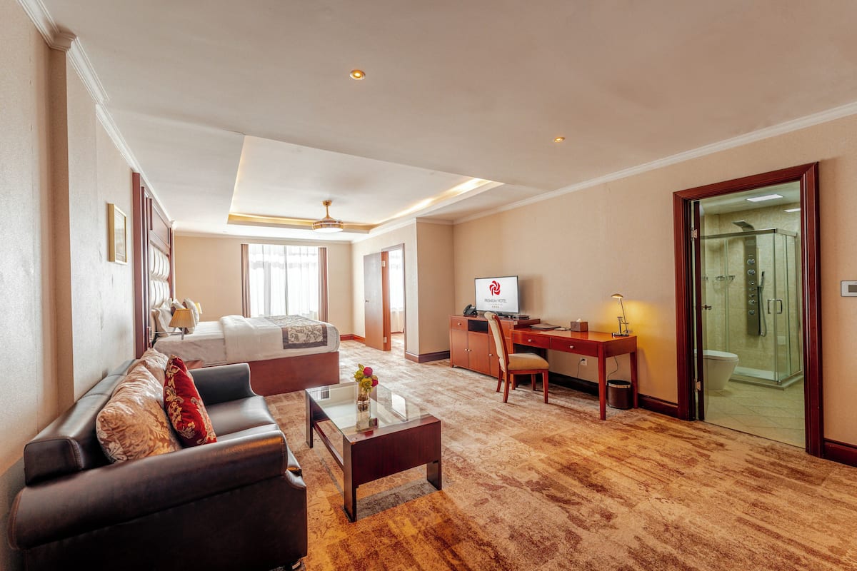 A hotel room with a living area featuring a sofa, a coffee table, and a TV. There is a desk with a chair, a glass shower enclosure, and a bed with a window.