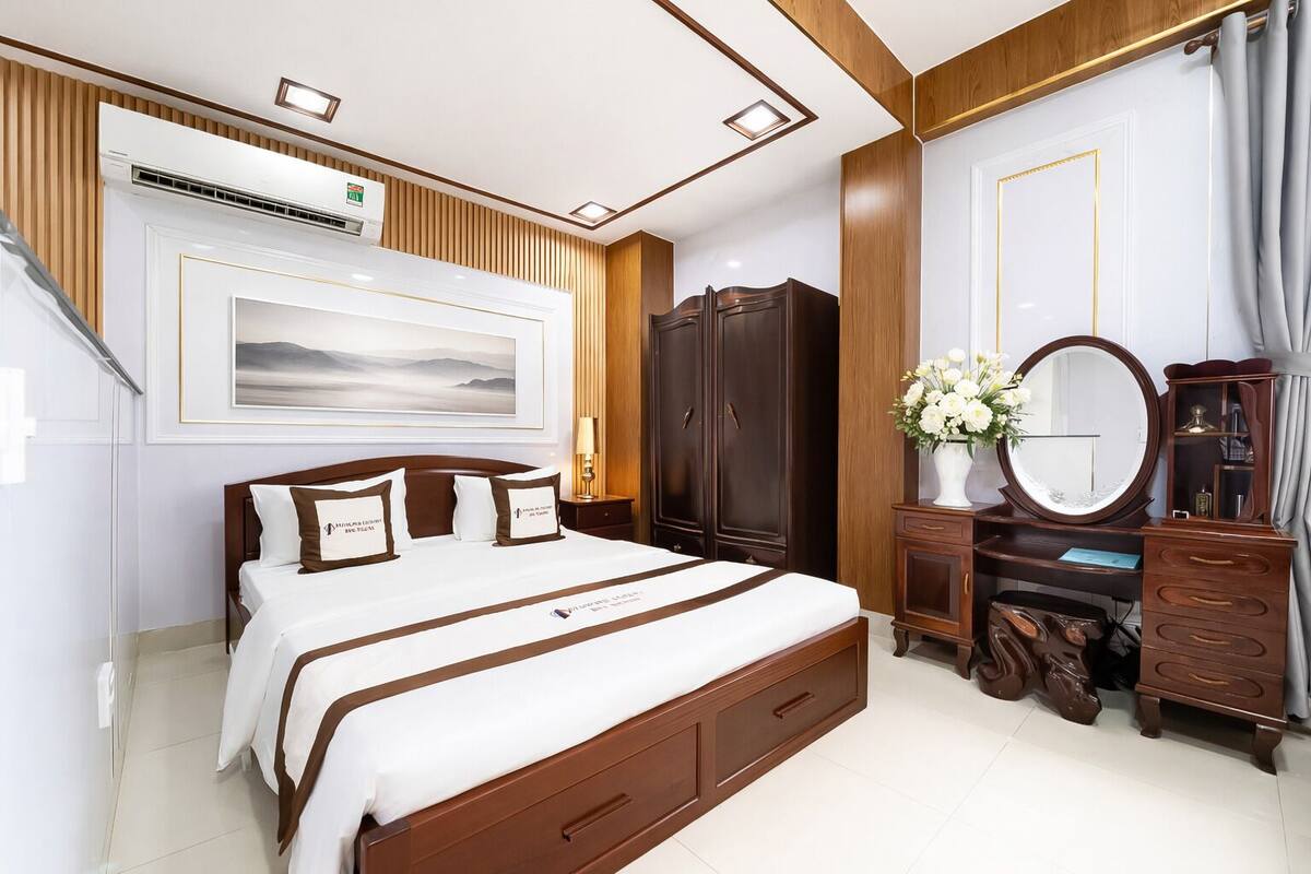 A hotel room with a large bed, wooden wardrobe, a vanity with a mirror, and a wall-mounted air conditioner.