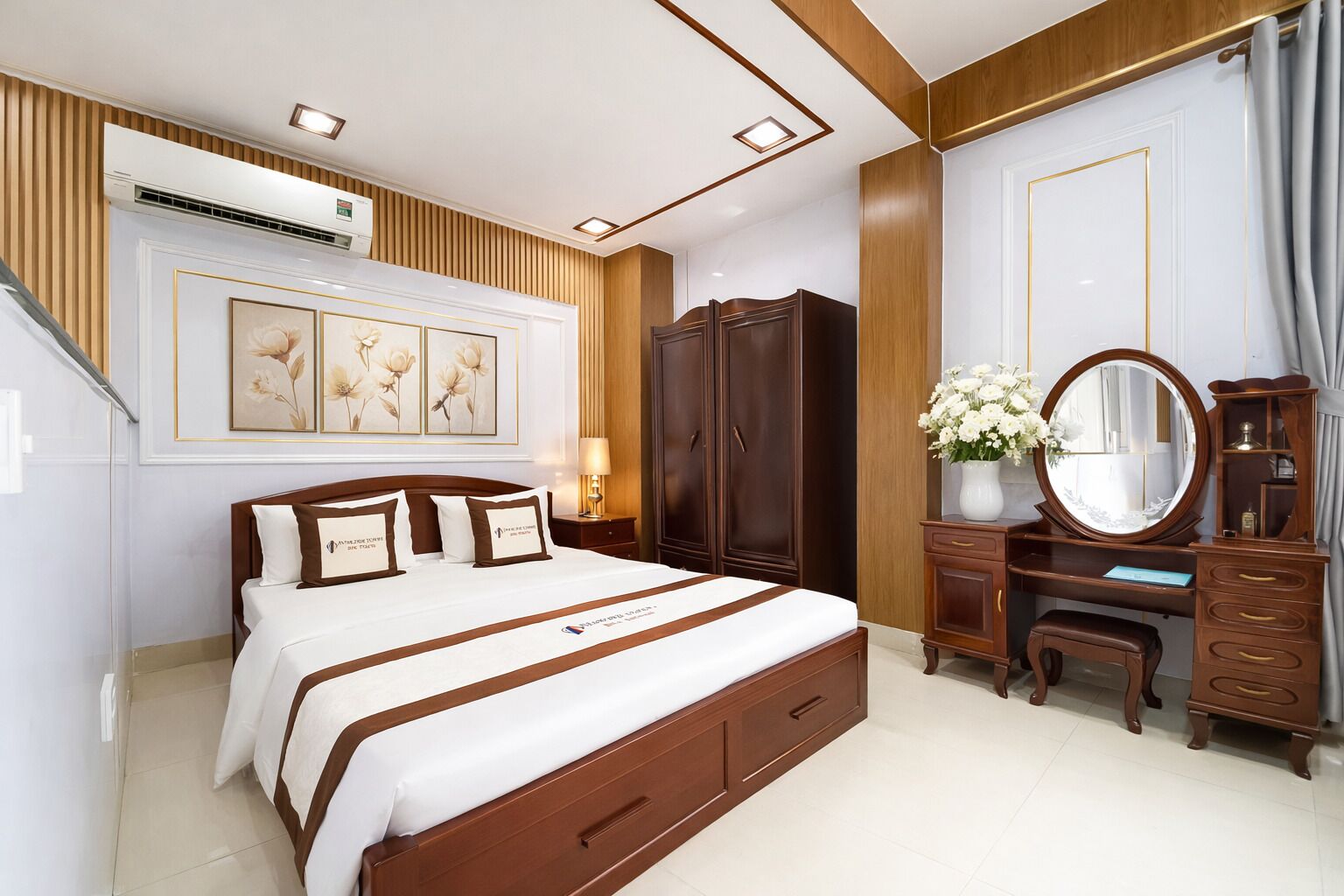 A hotel room with a large bed, wooden wardrobe, vanity with mirror, and wall-mounted air conditioning unit.