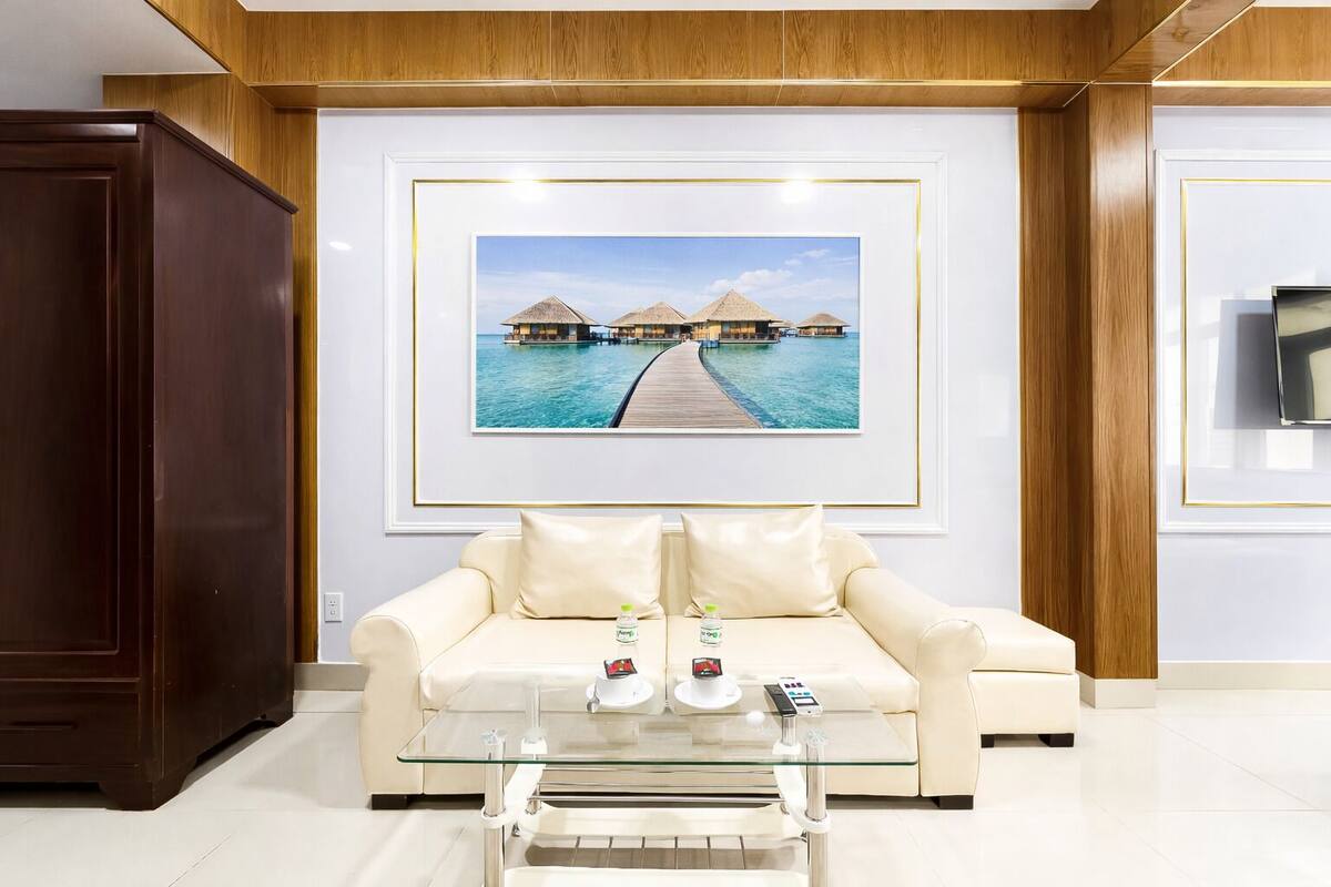 A modern living room with a beige sofa, a glass coffee table, and a framed picture of overwater bungalows.