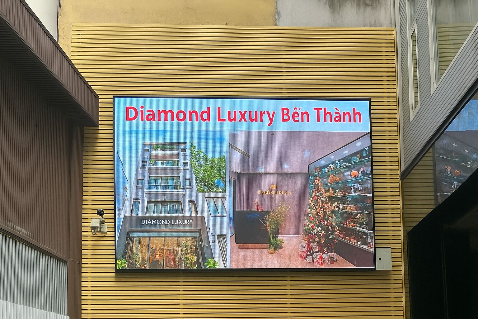 Un cartel publicitario de 'Diamond Luxury Bến Thạnh' muestra un edificio moderno y una exhibición de varios artículos.