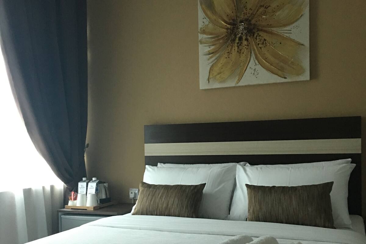 Un lit bien fait, avec du linge de lit blanc et deux oreillers marron, une œuvre d’art représentant une fleur accrochée au mur, et une table de chevet avec une machine à café et des tasses.