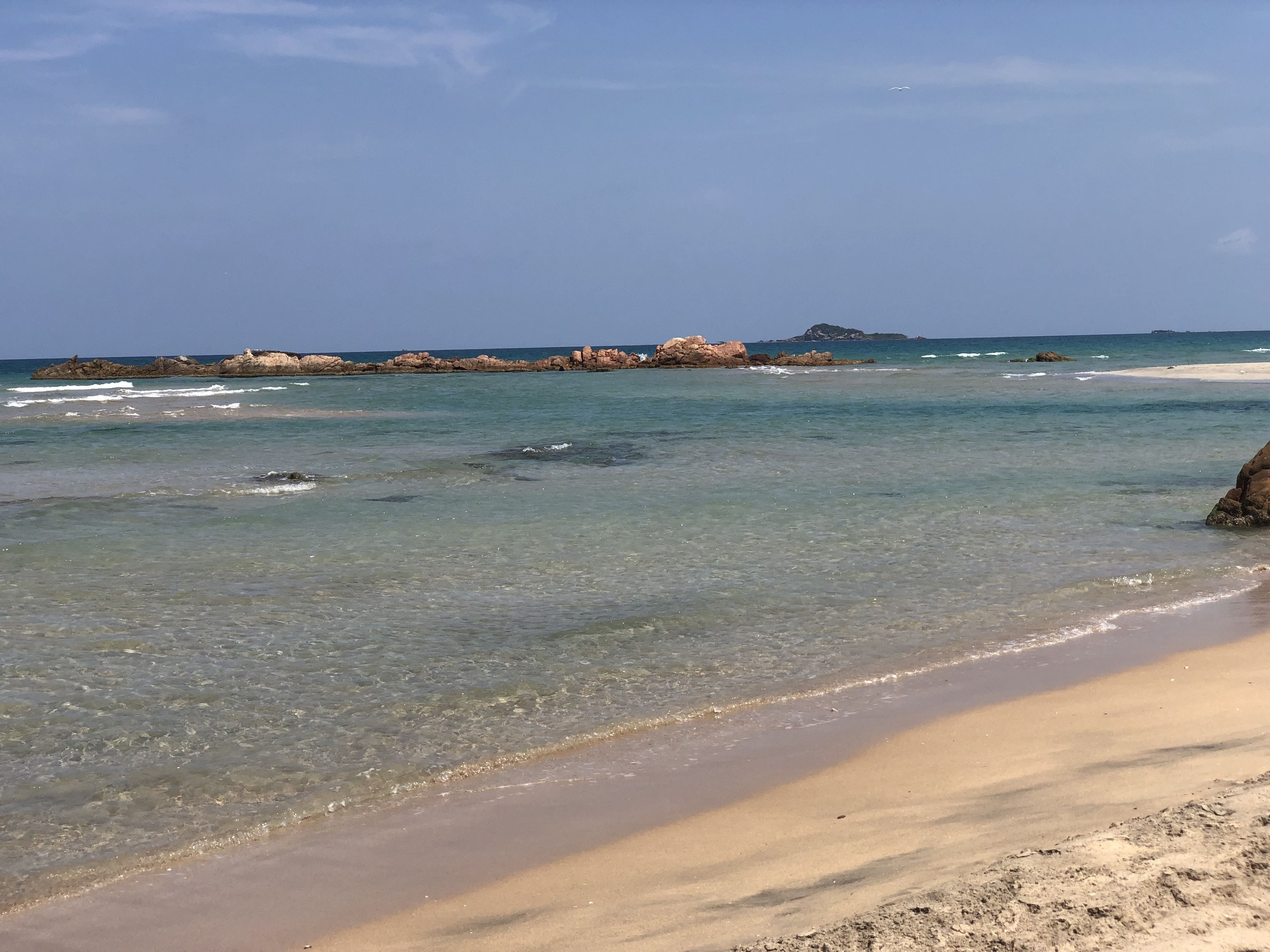 Una playa con aguas poco profundas y cristalinas, una orilla arenosa y formaciones rocosas a lo lejos.