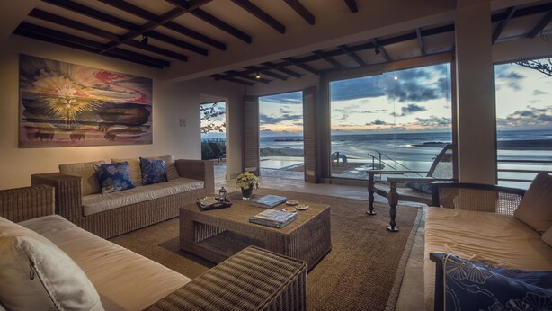 Amplia sala de estar con un gran ventanal con vista a la playa, con muebles de mimbre y un cuadro en la pared.