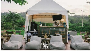 A tented outdoor seating area with a drink menu board, cushioned seating, and a bartender.