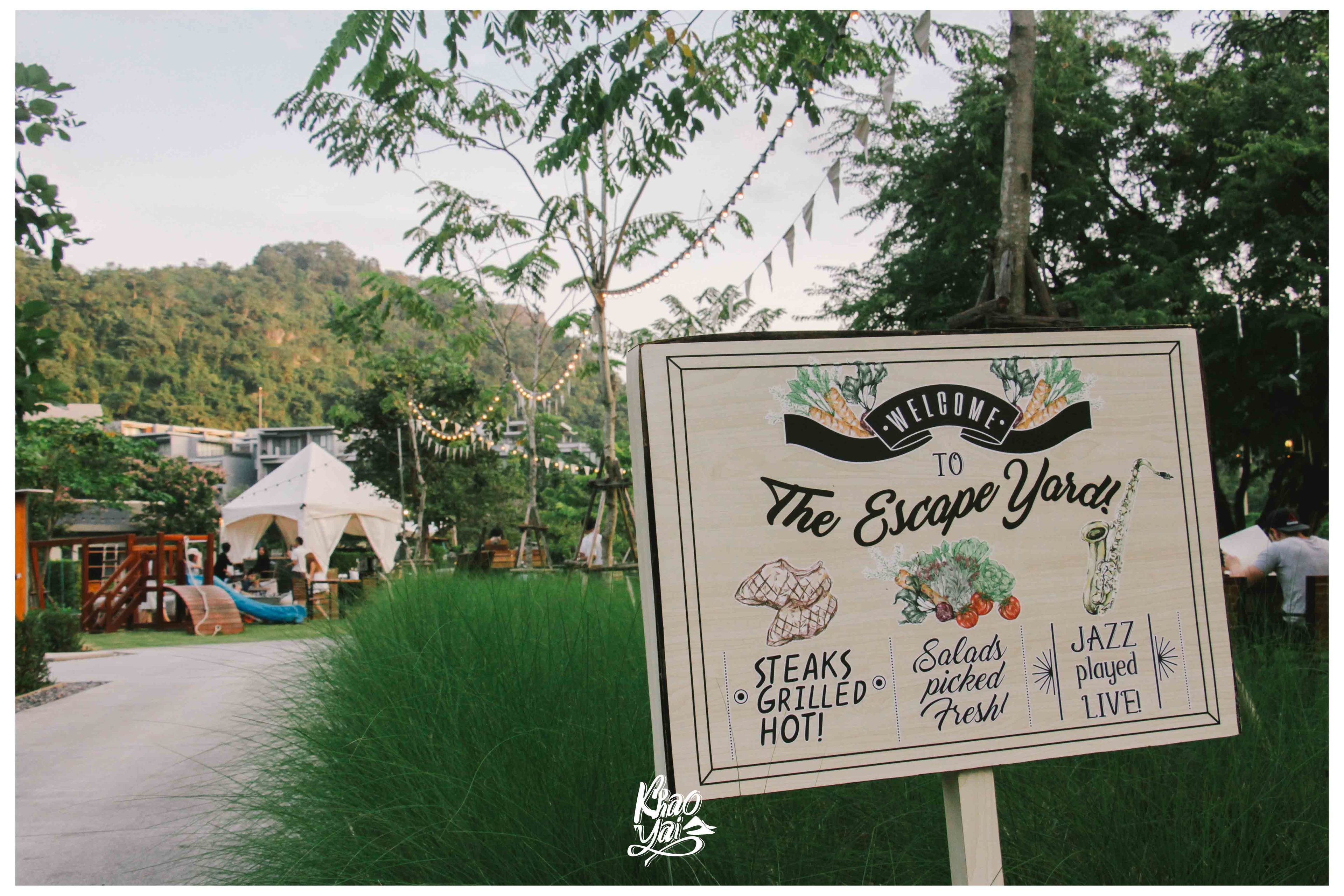 Un letrero da la bienvenida a visitantes a The Escape Yard, destacando opciones como filetes a la parrilla, ensaladas frescas y música jazz en vivo.