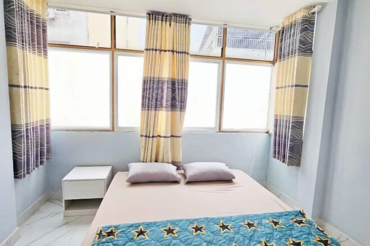A bedroom with a bed, two pillows, a bedside table, and a window with curtains.
