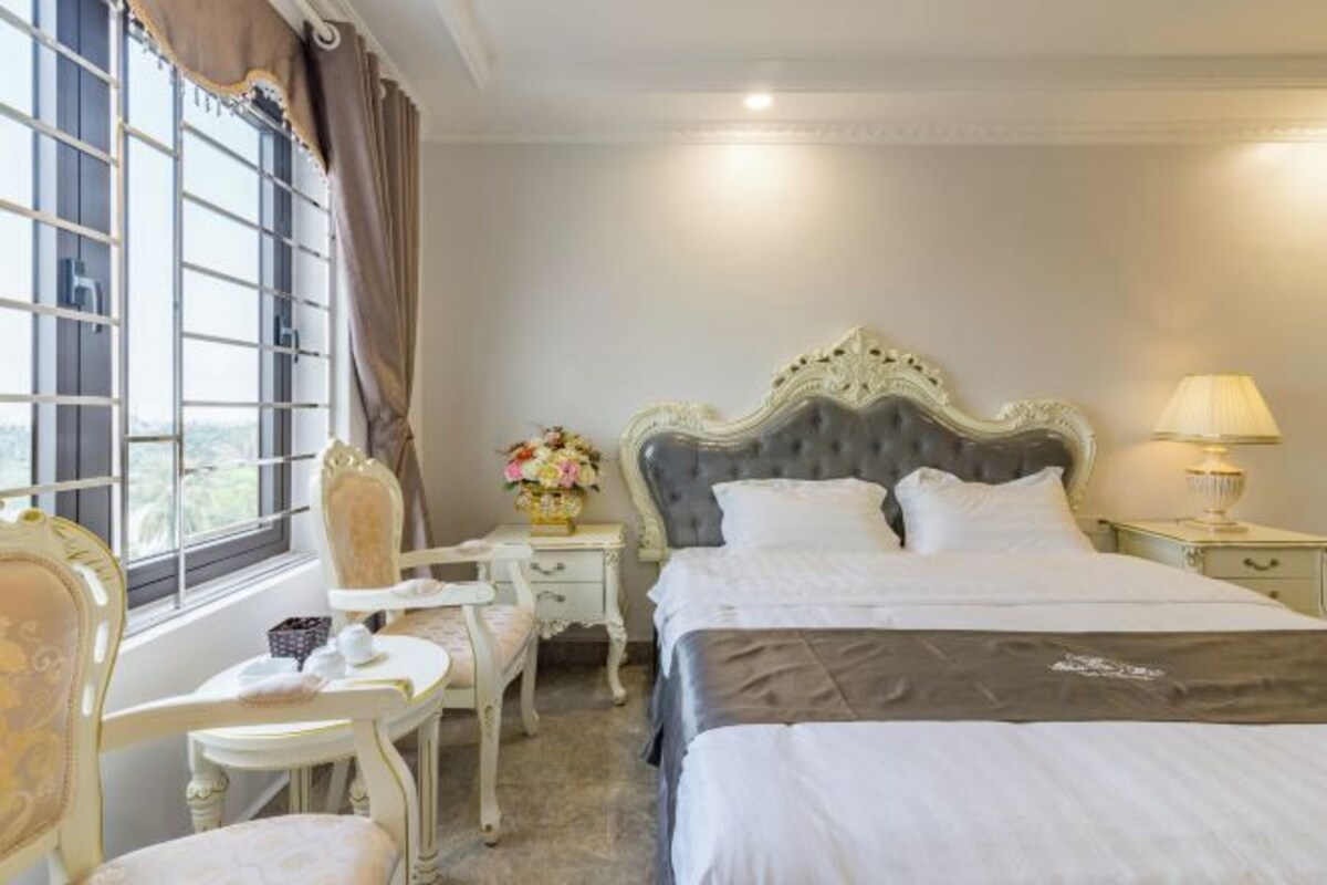 A bedroom with a large bed, a bedside table, a chair, a window with curtains, and a lamp.