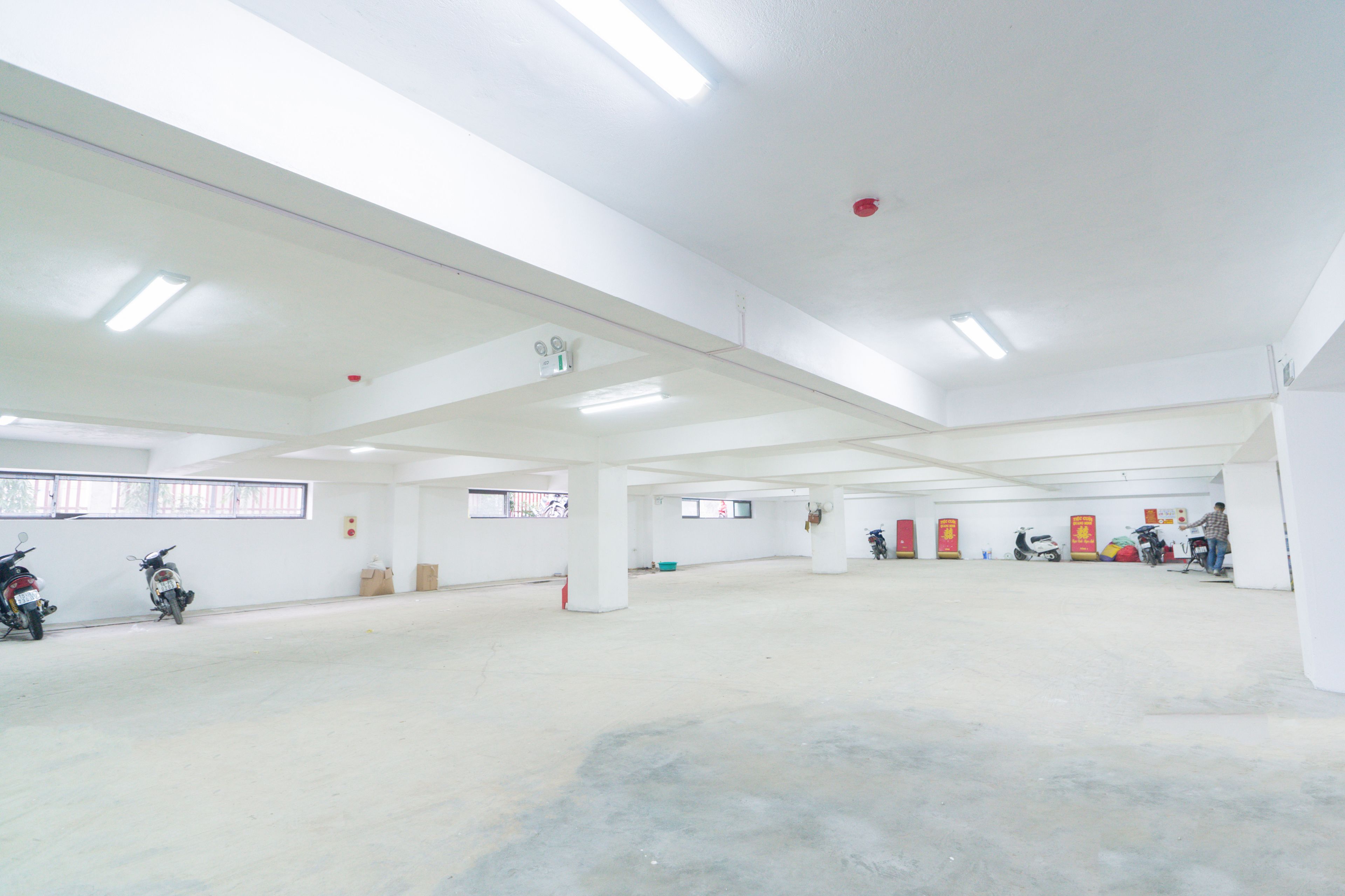 A spacious indoor area with white walls and ceiling, fluorescent lighting, and several motorcycles parked against the walls.